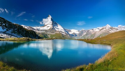 Naklejka premium Panorama of the Alps: Clear mountain lake with Matterhorn in the background, surrounded by lush greenery and snow-capped peaks under a blue sky