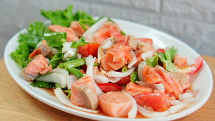 Vibrant Thai-style spicy salmon salad featuring fresh salmon chunks, onions tomatoes and herbs garnished with green vegetables and served on a white plate. 