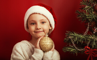Smiling cute kid girl holding in hands Christmas tree ornament golden ball near the holiday fur green tree in red santa claus hat. Happy expression winter holadays. Closeup