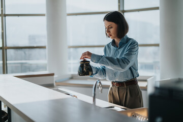 A focused woman prepares coffee in an open office kitchen with a modern, minimalist interior. The glass windows provide natural light, creating a peaceful and professional atmosphere.