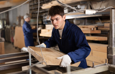 Young male carpenter putting wooden board on cutting machine in wood workshop