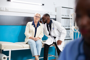Obraz premium Older white woman seated on hospital bed as african american doctor inspects her ear. Medical examination is being performed by skilled male physician using otoscope on senior female patient.