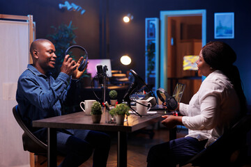 Two african content creators preparing to record online talk show with podcast equipment in home studio. Smiling male and female vloggers holding their headphones while talking on microphones.