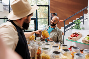Customer holds glass jar filled with organic food product and discusses with the seller wearing a hat and apron in eco friendly shop. Showcasing excellent customer service is a caucasian vendor.