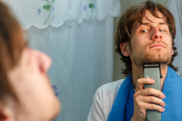 Young Man Trimming Beard with Electric Razor in Bathroom Mirror