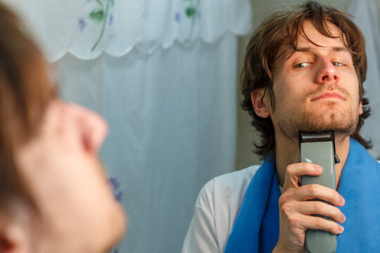 Young Man Trimming Beard with Electric Razor in Bathroom Mirror