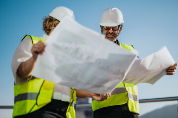 Two architects in safety vests and hard hats review building plans outdoors, symbolizing teamwork...