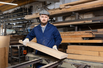 Male worker in uniform carries board from storage location to processing location. Selects planks for furniture manufacture