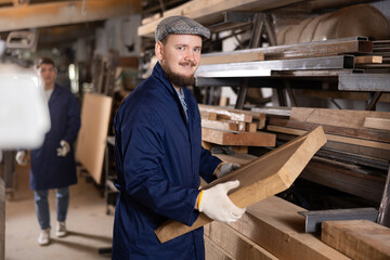 Male worker in uniform carries board from storage location to processing location. Selects planks for furniture manufacture