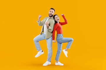 A joyful young couple stands back to back with arms raised in excitement. They express happiness and playfulness in front of a vibrant yellow backdrop, showcasing their close relationship.