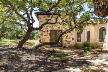 Obraz premium Rustic stone house surrounded by mature trees and lush landscape in daylight