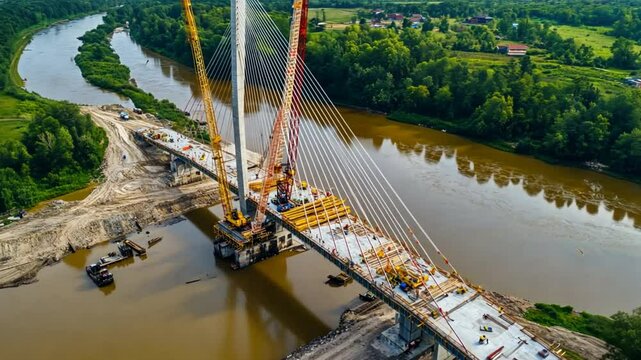 Aerial view of a cable-stayed bridge being built, with sections of the deck lifted into position by cranes. Workers secure each section as they connect across the span of the river.
