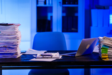 A quiet office desk at night, illuminated by a desk lamp. Documents, papers, and files are scattered across the desk, reflecting a late-night overtime work atmosphere.