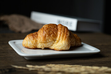 A golden-brown croissant, flaky and buttery, resting on a white plate against a dark background.