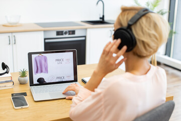 Happy Caucasian woman ordering clothes online using laptop while listening to music in headphones. Home setting includes modern kitchen background inspiring relaxed mood.