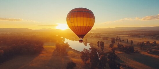 Obraz premium Hot air balloon flying over a river and forest at sunrise with the sun shining through the clouds.