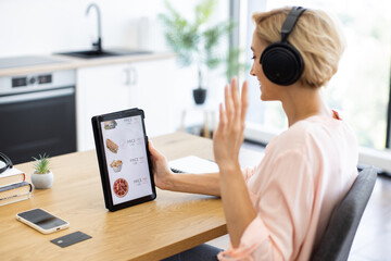 Happy Caucasian woman using tablet to order food online while enjoying music in headphones. Relaxed atmosphere with modern kitchen background, highlighting ease of online shopping at home.