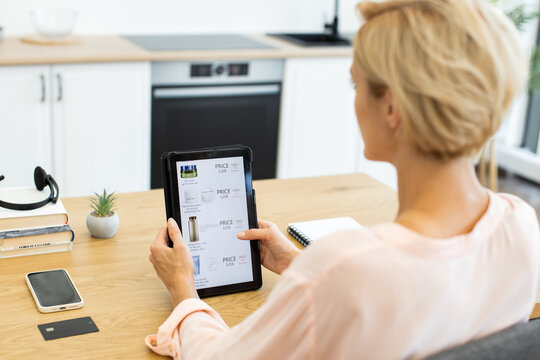 Middle-aged Caucasian woman using tablet to order cosmetics online in modern kitchen. Image shows convenience of online shopping, with focus on digital technology and home lifestyle.