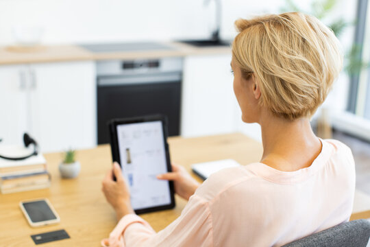 Caucasian woman browsing online shopping website on tablet in modern kitchen. Middle-aged female checking out latest cosmetics available for delivery.
