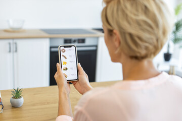 Caucasian woman using smartphone to order food online through delivery website. She is seated in modern kitchen, browsing various food options on mobile device.