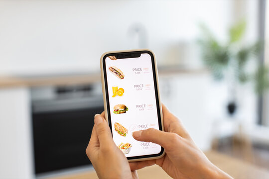 Caucasian woman using smartphone for online food order in bright modern kitchen. Person browsing delivery app menu with various meal options like sandwiches and juices.