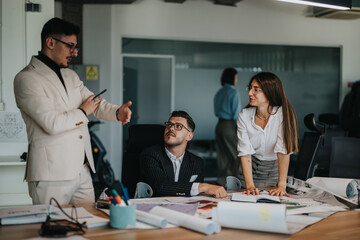 A project leader on crutches discusses paperwork with team members at a table filled with documents and plans in an office environment.