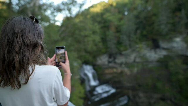 woman with long hair capturing photo of waterfall using her smartphone, surrounded by lush greenery in tranquil forest setting at Cummins Falls State Park, USA. woman capturing photo of waterfall