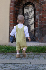 Preschool boy in ochre romper and flat cap happily back. Child walks on old cobblestones in city...