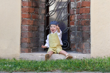 Preschool boy in ochre romper and flat cap pointing index finger up joyfully. Child sitting happily...