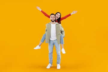 A young couple joyfully embraces their relationship as the woman playfully rides on her partner's back. They showcase happiness and connection, set against a bright yellow background.