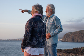 Back view of senior couple standing at sunset light on the rocks at sea looking at the horizon over water. Relaxed lifestyle for a caucasian couple of retirees enjoying travel and nature