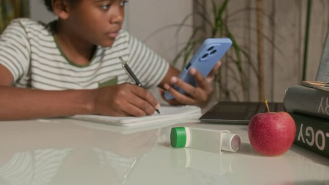Young African American boy holding smartphone and writing notes in copybook while doing homework, textbooks, apple and asthma inhaler on table nearby in foreground