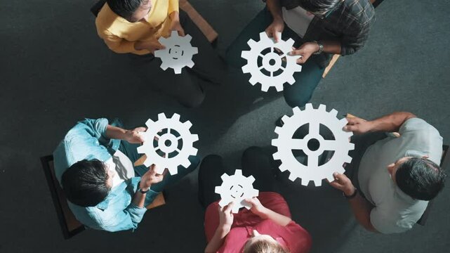 Top view of smart business people joining and gathering white gear together. Aerial view of diverse team working together to put or collect cog while sitting in circle. Unity, team work. Symposium.