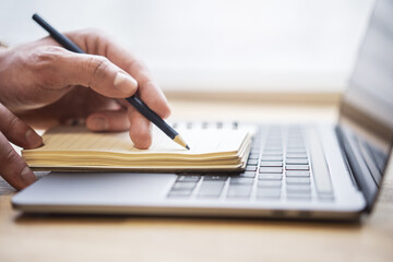 The image highlights a man's hand writing in a notepad on a laptop, with a faintly blurred...