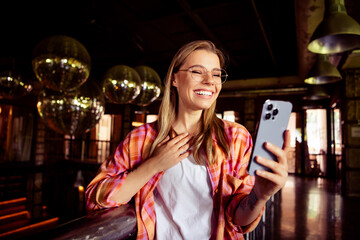 Photo of lovely young blonde lady video call device laugh dressed plaid shirt comfortable startup dark coworking office dark cafe interior