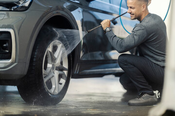 Happy man is kneeling and using high pressure water jet gun for washing car tires at self service...