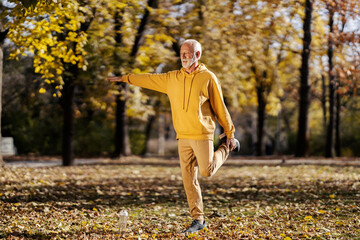 An old fit athlete in tracksuit balancing on one foot and stretching it while preparing for fitness...