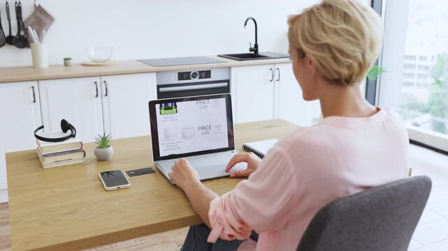 Caucasian woman using laptop to browse cosmetics on delivery website. Middle-aged female shopping online at home, sitting in modern kitchen.
