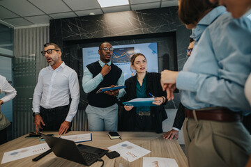 A multicultural group of business people actively discussing at a meeting table in a modern office setting. Charts and laptops indicate a focus on business strategy and collaboration.