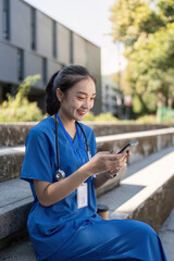 Asian nurse texting outdoors in blue scrubs