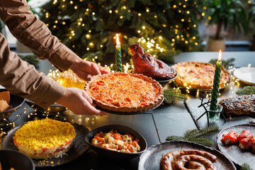 Man places Christmas quiche pie on festive table surrounded by many dishes in cozy evening atmosphere with Christmas tree and garlands