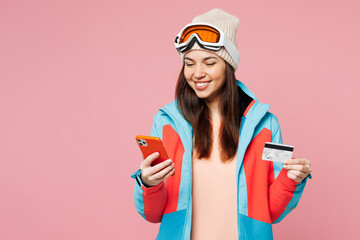 Skier young woman wear blue padded jacket ski goggles mask spend extreme weekend winter season in mountains hold use mobile cell phone credit bank card isolated on plain pink background Hobby concept. © ViDi Studio