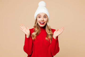 Merry shocked surprised young woman wears red warm cozy knitted sweater hat posing spread hands isolated on plain pastel light beige background. Happy New Year celebration Christmas holiday concept.