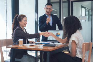 Successful Partnership: Businesswomen shaking hands, celebrating a deal with a cheerful businessman in the background. A modern office setting enhances the professional atmosphere. 