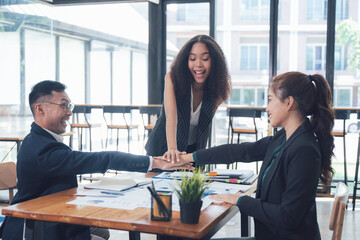 United in Success: A diverse team of business professionals in a modern office setting, their hands stacked together in a gesture of unity and collaboration. They are excited and happy.