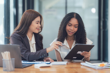 Collaborative Success: Two businesswomen, representing different ethnicities, engage in a productive discussion, reviewing data on a tablet.