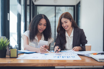 Collaborative Businesswomen Analyzing Data: Two smiling businesswomen, one African American and one Asian, collaborate closely, reviewing financial charts and graphs.