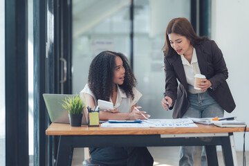 Collaborative Workspace: Two diverse businesswomen engage in a productive discussion, reviewing documents and ideas together in a modern office setting.
