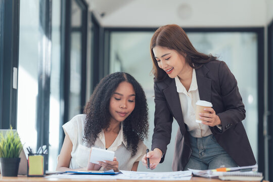 Collaborative Spirit: Two diverse businesswomen, one with a coffee cup in hand, engage in a productive discussion over blueprints.