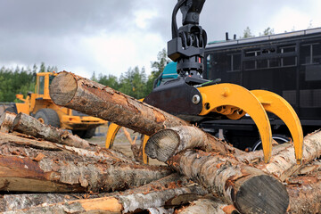 Log loader grapple attachment unloads wooden logs at a work site with heavy machinery on the background.